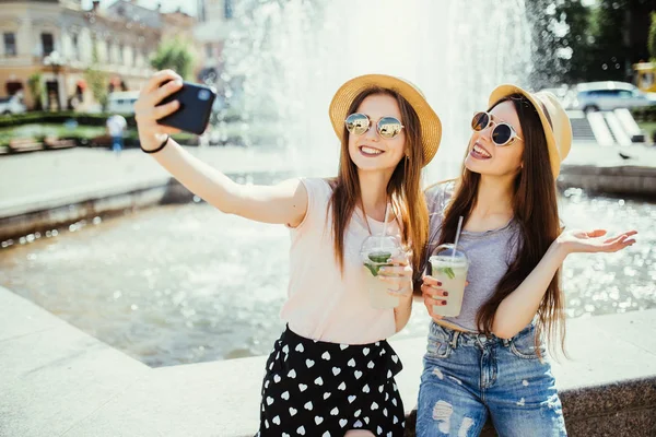 两个开朗的女人对喷泉与冰沙 使自拍在现代手机或在线聊天视频通话 分享他们对国外度假胜地的印象 — 图库照片
