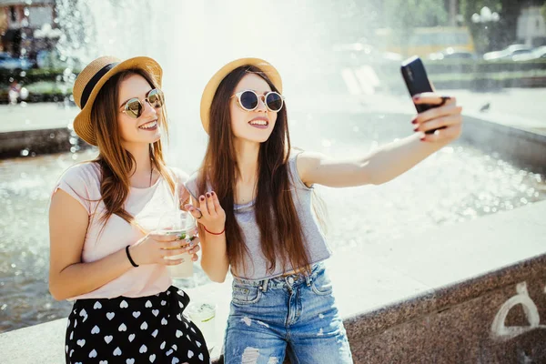 happy young two friends women taking selfie by smartphone and drinking cocktails drinks outdoor in the summer street