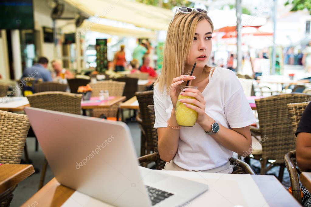 Beautiful young woman sitting outdoors and chatting, drinking cocktail using laptop a in cafe.