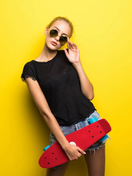 时尚快乐微笑时髦酷的女孩在太阳镜和五颜六色的衣服与滑板有乐趣户外对橙色背景 — 图库照片
