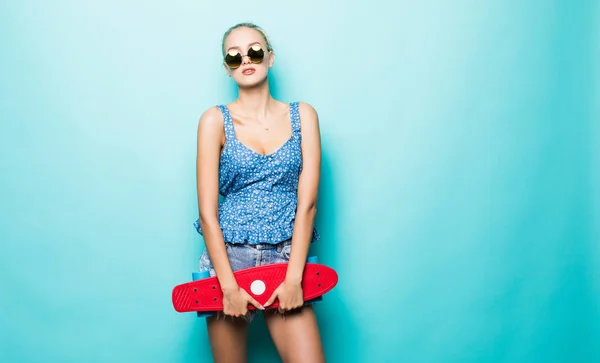 有吸引力的女孩拿着滑板在帽子和太阳镜摆在蓝色背景上 — 图库照片
