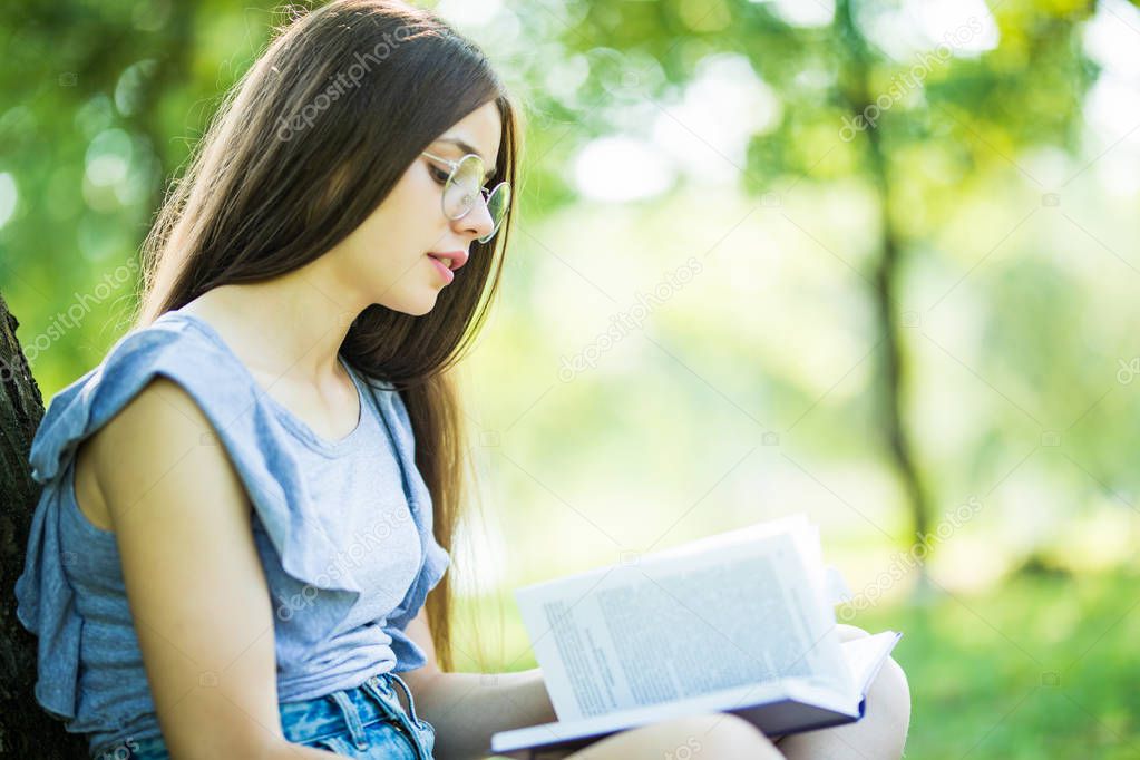 Side view of pleased woman in eyeglasses sitting on the grass under tree and reading book in park
