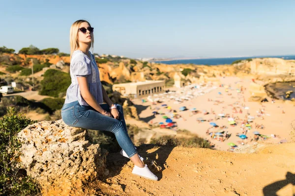 Young woman wear in sunglasses and wear casual clothes sitting on hight stone and enjoying the view on the beach