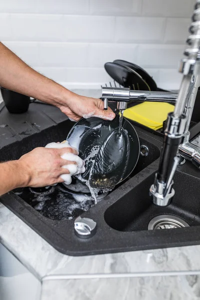 The man washes the dirty dishes in the kitchen by hand. The man holds a dirty plate and a washing sponge in his hand. The concept of washing dirty dishes by hand.