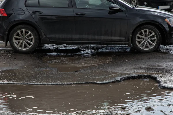 The car goes without reducing speed on a bad road with asphalt in pits and potholes, hitting the wheel in a puddle, spraying melt water in all directions — Stock Photo, Image