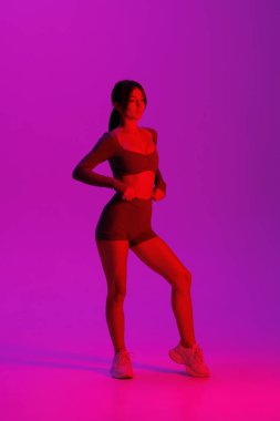 Spor fitness kadını, uzun saçlı, arkası bağlı, pürüzsüz mor bir giysi giyiyor, yumuşak bir vücut, hafif ışıklandırma, modern spor kıyafeti, sağlıklı bir yaşam tarzı.