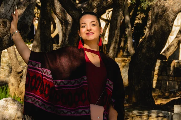 Yunanistan 'ın Akropolis kentinde Herodes Atticus Odeon' un ağaçlarının yanında poz veren panço ve kırmızı küpeler takan gülümseyen kadın. Tarihi eser üzerinde seyahat portresi.