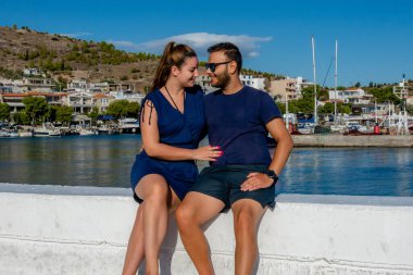 Pachi, Megara, Yunanistan limanında birlikte oturan çift. Arka planda yatlar, tekneler ve sahil kasabasıyla yaz manzarasının keyfini çıkarıyor ve gülümsüyor.