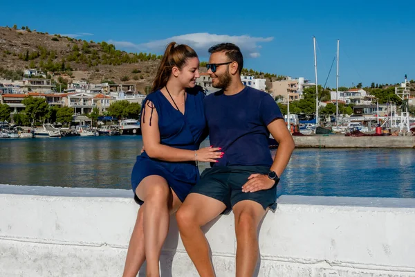 Pachi, Megara, Yunanistan limanında birlikte oturan çift. Arka planda yatlar, tekneler ve sahil kasabasıyla yaz manzarasının keyfini çıkarıyor ve gülümsüyor.