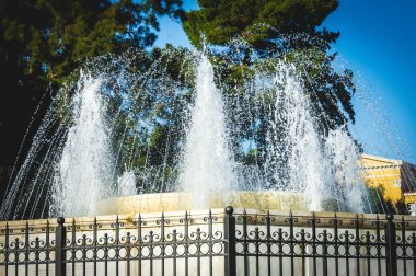 Yunanistan 'ın Atina kentindeki Zappeion Hall yakınlarında etrafı demir çitlerle çevrili güçlü su jetleriyle çevrili mermer bir çeşme ağaçlara ve açık mavi gökyüzüne karşı kurulmuş..
