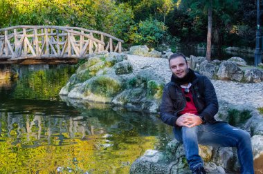 Yunanistan 'ın Atina Ulusal Bahçesi' ndeki gölet ve ahşap köprü kenarında poz veren genç bir adam, tarihi bir halk parkı ve şehir merkezinde doğal bir simgeyi rahatlatıyor..