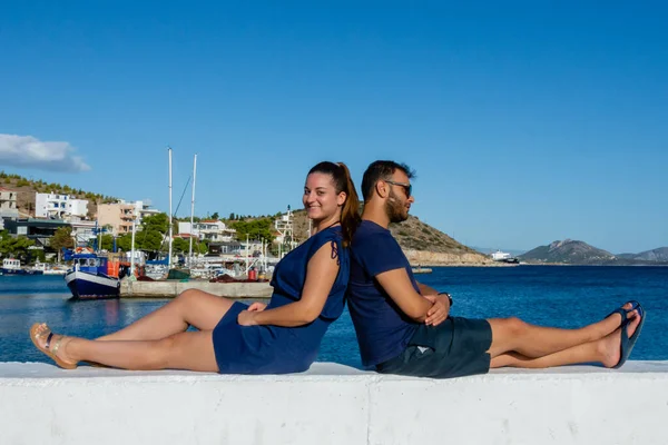 Yunanistan 'ın Pachi kentindeki rıhtımda dinlenen genç çift, arka planda marina, balıkçı tekneleri, yatlar ve tepelerle beyaz duvarda sırt sırta oturuyorlar..