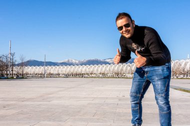 Atina, Yunanistan 'daki Olimpiyat Spor Kompleksi' nin Santiago Calatrava çatısında baş parmağıyla poz veren mutlu adam, arka planda dağlarla birlikte.