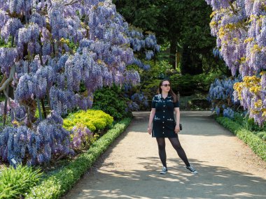 Genç ve güzel bir kadın, çiçeklenmiş mor salkımın yanında zarif bir şekilde duruyor..