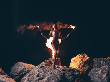 Bikini muhteşem bir ince vücut ile genç seksi kadın elinde ateş meşaleleri ile yanan kaya üzerinde karanlık gece (poz) kalıyor. Arka planda siyah gökyüzü