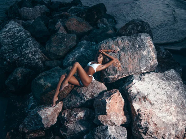 ビキニでゴージャスなスリムな体を持つエレガントな 見事な若い女性は 夜に岩の上に横たわってポーズをとっています 海岸で夏の暗い屋外写真撮影 海の近くのビーチでの写真 — ストック写真