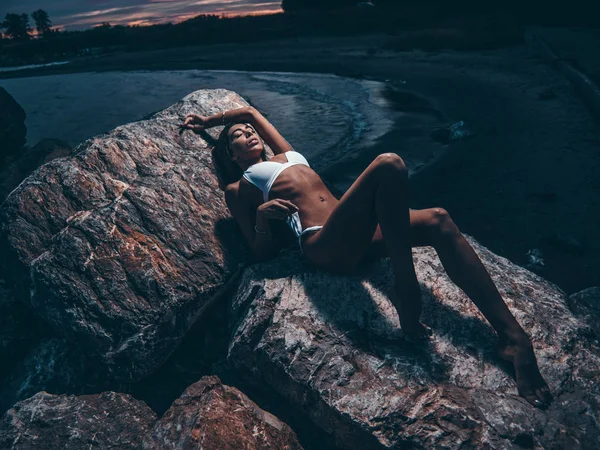 水着姿の魅惑的なスリムな体を持つ官能的でセクシーな若い女の子が 夜に岩の上に横たわってポーズをとっています 夏の暗い屋外の写真撮影は 夕暮れ時の海の近くのビーチでの写真 — ストック写真