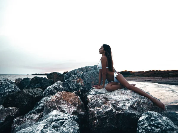 ビキニで魅力的なスリムな体を持つエレガントでセクシーな若い女性は 夕日に岩の上に座ってポーズをとっています 海岸で夏の屋外写真撮影 水の近くのビーチでの写真 — ストック写真