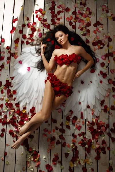 彼女の背中の後ろに長いブルネットの緩い髪と白い翼を持つ美しい 壮大な裸の若い女性は 天使としてスタジオで横たわってポーズをとっています 床には赤いバラの花びらがたっぷり キューピッド — ストック写真