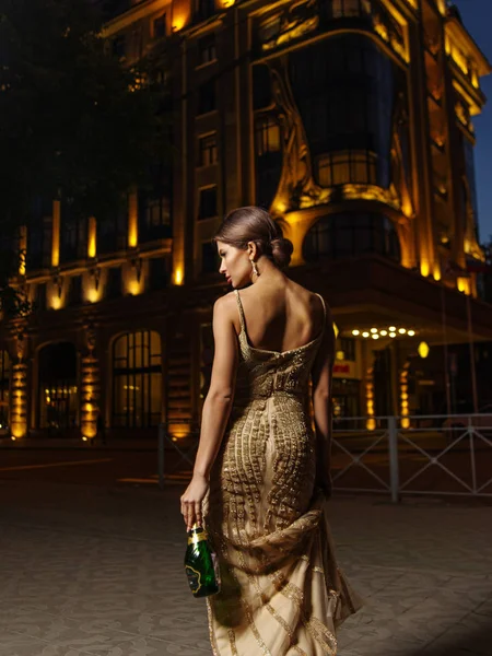 Elegant rich smartly dressed woman with beautiful hairstyle wearing expressive sparkling evening golden dress is walking to the street at night holding an expressive bottle of champagne. Back view