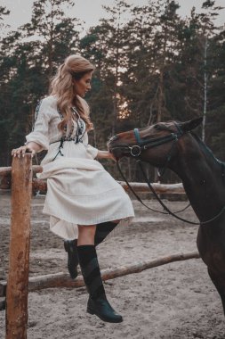 Elbise giyen güzel ve zarif genç kadın çiftlikte at okşayarak. Çekici bir binici eyer yakınında açık havada poz veriyor. Yaz, doğa manzarası, kırsal