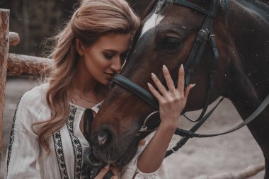 Elbise giyen güzel ve romantik genç kadın kucaklayan ve çiftlikte at okşayarak. Çekici bir binici eyer yakınında açık havada poz veriyor. Yaz, doğa manzarası, kırsal