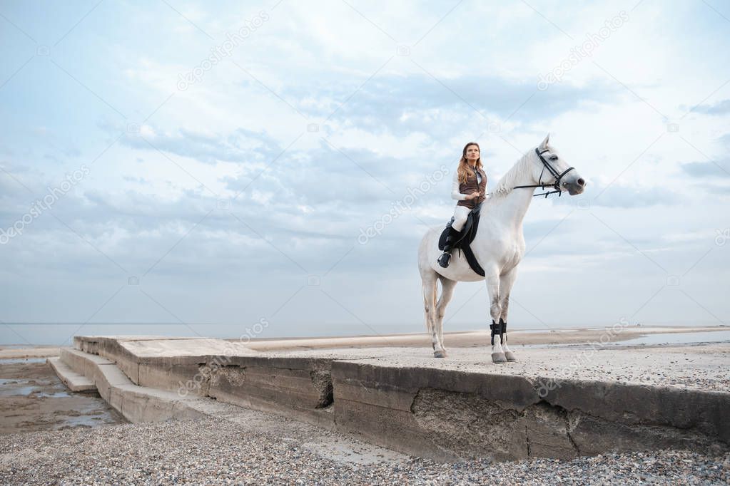 Elegant and beautiful confident young woman wearing stylish jockey outfit is holding reins and riding a white horse on the coast. An attractive rider is posing outdoors.Summertime, nature landscape