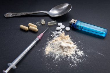 Tıbbi bir şırınga, bir kaşık, bir çakmak ve siyah arka planda beyaz unla karıştırılmış ezilmiş tıbbi tabletler. Yasadışı uyuşturucu kullanımı, bağımlılık konsepti.