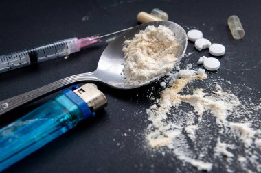 Tıbbi bir şırınga, üzerinde beyaz un olan bir kaşık, bir çakmak ve siyah arka planda ezilmiş tıbbi tabletler. Yasadışı uyuşturucu kullanımı, bağımlılık konsepti.