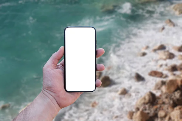 Guy holds in his hand a smartphone close-up, with a white screen on a background of sea. Mock-up Technology