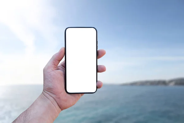 男は海の背景に白い画面で 彼の手にスマートフォンのクローズアップを保持しています モックアップ技術 — ストック写真