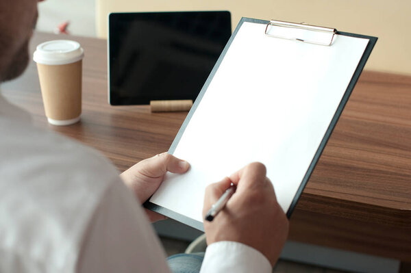 Бизнесмен в белой рубашке с цифровым планшетом в руках подписывает контракт в офисе. Рабочее место с чашкой кофе и документом с ручкой на деревянном столе