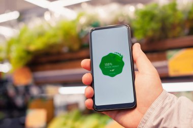 Doğal ürün. Bir adam elinde elinde bir akıllı telefon, ekranında bir etiketle süpermarketteki bir vitrinde taze yeşilliklerin arka planına karşı duruyor. Çevre dostu ve sağlıklı yiyecekler.