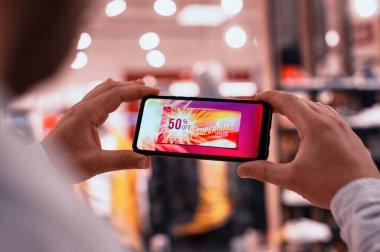 Bir adam elinde bir akıllı telefon tutuyor ve ekranında bir yazıyla bir giyim mağazasının arka planında Süper Satış yapıyor.