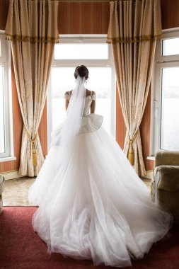 Gelinlik giymiş gelin pencerenin yanında duruyor ve elbiseyi omuzlarının üzerine indirdi. Harika ışık..