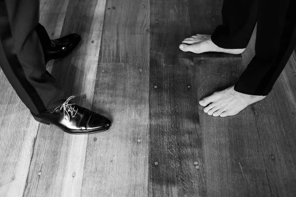 Two men in pants. One is wearing shoes, the other is barefoot. Standing on a wooden floor.