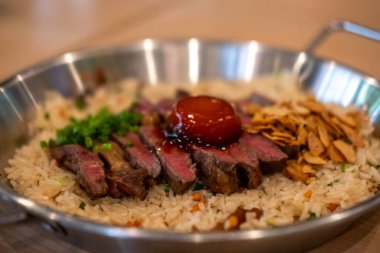 Sarımsaklı kızarmış pirinçli biftek, üzerine az pişmiş sığır eti ve tavada balık soslu yumurta sarısı.