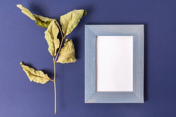 An image of a blue wooden frame. Watercolor paper inside the frame with place for text. Blue-purple background made of pastel embossed paper. Dried large leaf to the right of the frame. View from above.