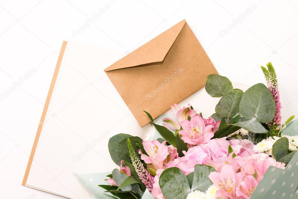 Open vintage craft notebook or diary with tender flowers decoration and closed craft envelope on the white table. Floral mock-up and empty space for text. Bouquet of pink flowers. Flat lay.