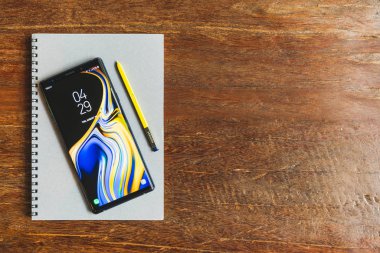 Bangkok, Thailand - Aug 30, 2018: Ocean Blue Samsung Galaxy Note 9 with yellow S pen stylus on a notebook with copy space on wooden table. Illustrative Editorial. clipart