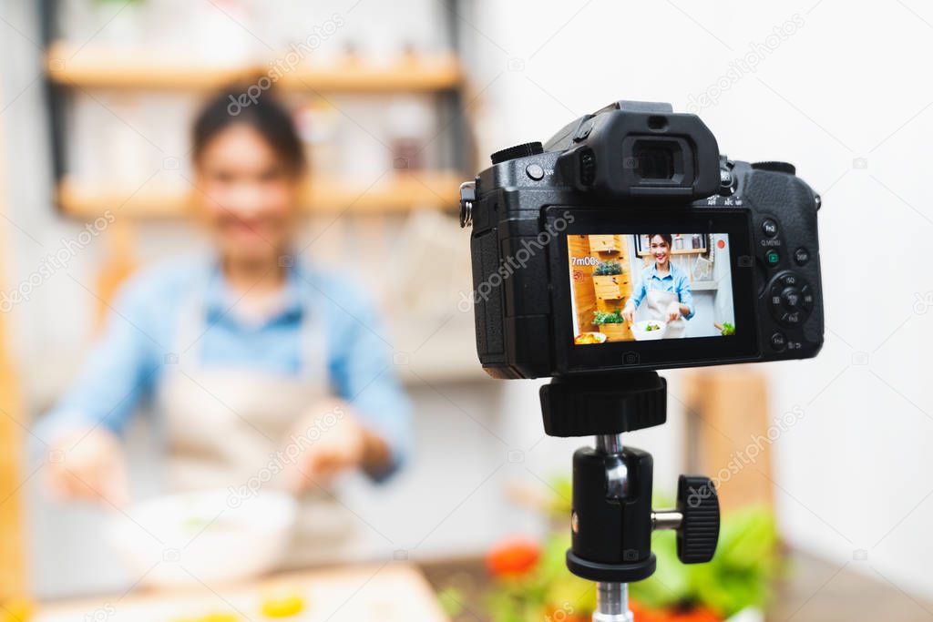 Young cute Asian blogger girl recording video tutorial session of salad cooking lesson at home kitchen. Food blogging or vlogging, social media hobby broadcasting, or online learning course concept
