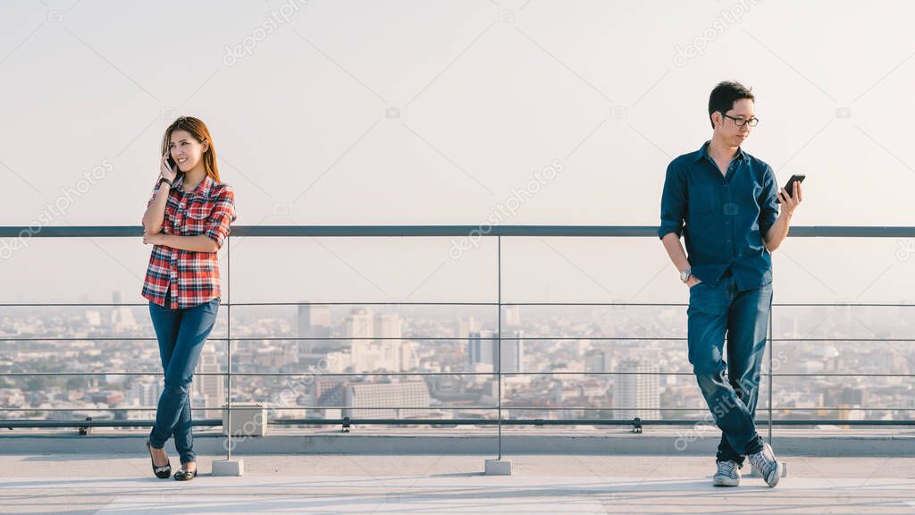 Young adult Asian couple using smartphone in distance on building rooftop. Coronavirus outbreak social distancing, new normal people lifestyle, or mobile phone telecommunication technology concept