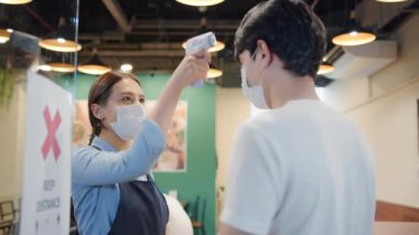 Asyalı bayan restoran çalışanları içeri girmeden önce erkek müşterinin alnına kızılötesi termometre veya ısı silahı kullanıyor. Coronavirus Covid-19 önlem ateşi kontrolü, yeni normal yaşam tarzı