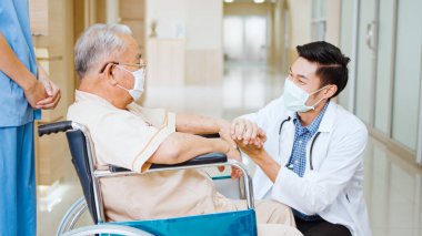 Genç Asyalı doktor el ele tutuşup hastane koridorunda tekerlekli sandalyedeki yaşlı hastayla konuşuyor. Sağlık hizmeti işi ya da hastane iş konsepti