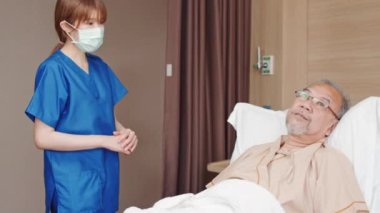 Bayan Asyalı hemşire maske takıyor ve hastane yatağında yatan son sınıf erkek gülümseme hastasını kontrol ediyor. Huzurevi, sağlık hizmeti, konukseverlik ya da tedavi konsepti