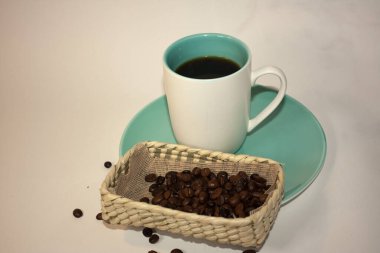 Kahve çekirdekleri mavi tabakta beyaz bir kahve fincanının yanında doğal bir lif kabının içinde.