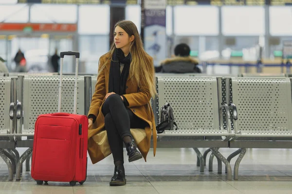 Airport woman waiting in terminal. Air travel concept with young casual business woman sitting with carry-on hand red bag.