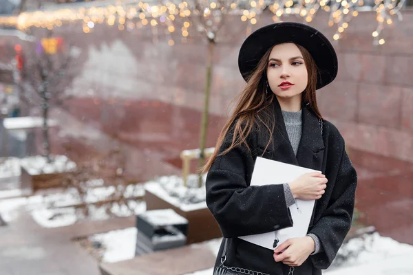 年轻的时尚女性拿着她的杂志 走在城市街道上戴着黑色的帽子环顾四周 对着镜头微笑 复制空间 图片仅供参考 — 图库照片