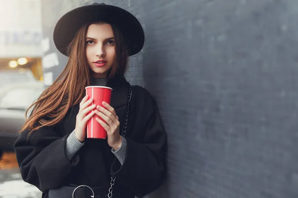 年轻的时尚女性拿着她热的一杯咖啡 站在墙边戴着黑色的帽子环顾四周 对着镜头微笑 复制空间 图片仅供参考 — 图库照片