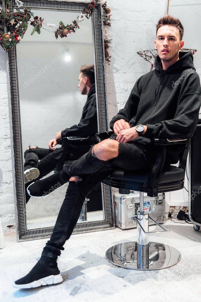 Fashionable man sit at hair salon with cup of tea. Wear blue jacket and all black. Winter, autumn outfit. Jacket with blouse and black sneakers.